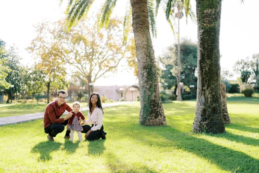 Smiling parents squatting near little girl on sunny meadow in park. High quality photo