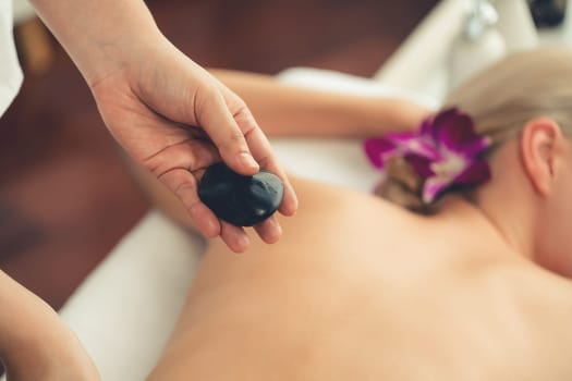 Hot stone massage at spa salon in luxury resort with day light serenity ambient, blissful woman customer enjoying spa basalt stone massage glide over body with soothing warmth. Quiescent