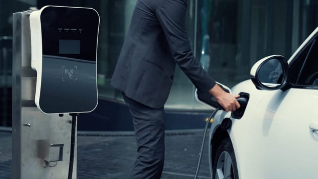 Progressive businesswoman install charger plug from charging station to his electric car before driving around city center. Eco friendly rechargeable car powered by sustainable and clean energy.