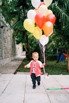 Little girl with a bunch of colorful balloons walks through a green garden. High quality photo