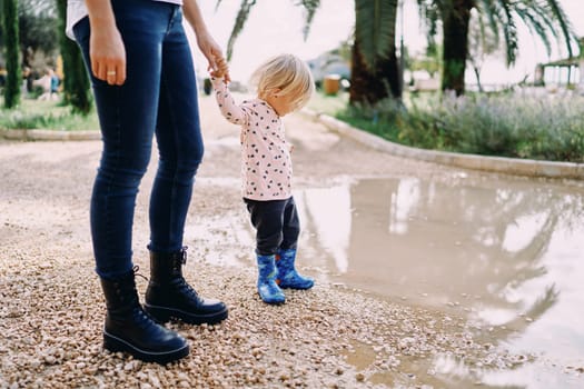 Mom holds the hand of a little girl in rubber boots standing on the edge of a large puddle. High quality photo