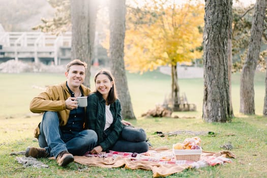 Smiling man with mug hugging woman while sitting on blanket in park near food basket. High quality photo
