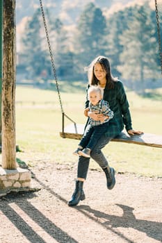 Little girl sitting on her mother lap swinging on a wooden swing in the park. High quality photo