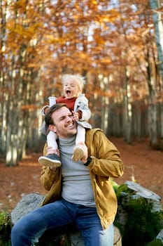 Little smiling girl sits on her dad shoulders touching his face with her hands in the autumn forest. High quality photo
