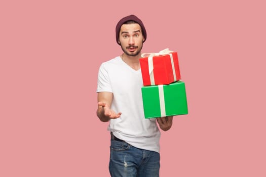 Portrait of puzzled confused bearded man in white T-shirt and beany hat holding two present boxes in hands, looking at camera with surprised expression. Indoor studio shot isolated on pink background