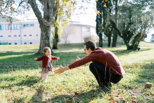 Little girl runs to her dad, who opened his arms in an embrace, squatting on the lawn. High quality photo
