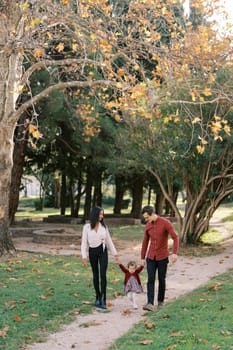 Mom and dad with a little girl walk along the path in the park, holding hands. High quality photo