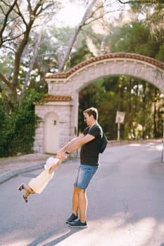 Smiling dad circling a little girl holding her hands on the road at the park gate. High quality photo