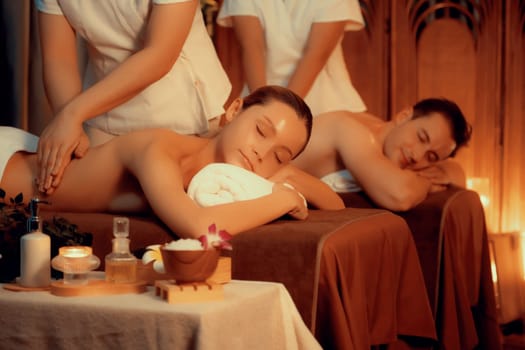 Caucasian couple customer enjoying relaxing anti-stress spa massage and pampering with beauty skin recreation leisure in warm candle lighting ambient salon spa at luxury resort or hotel. Quiescent