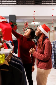 Employee presenting clothes to customer, asian woman looking for fashion items to give as christmas presents in retail store. to Shopping manager recommending accessories for dinner preparations.