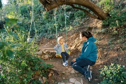 Mom swings a little girl on a rope swing sitting on the ground nearby. High quality photo