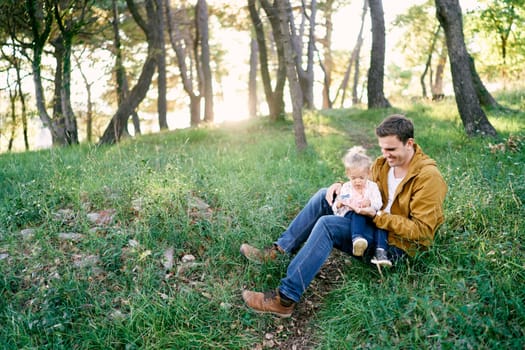 Dad with a little girl on his knees sits on a lawn in the forest. High quality photo