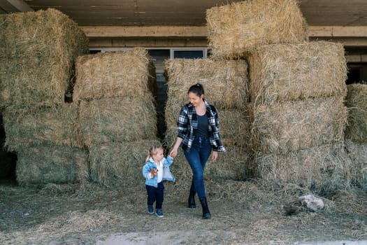 Mom and little girl walk through the hayloft past stacked bales of hay. High quality photo