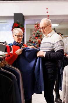 Senior people looking for blazers on racks, buying clothes on discount from clothing store at shopping center. Clients checking trendy items, searching festive attire and gifts for christmas.