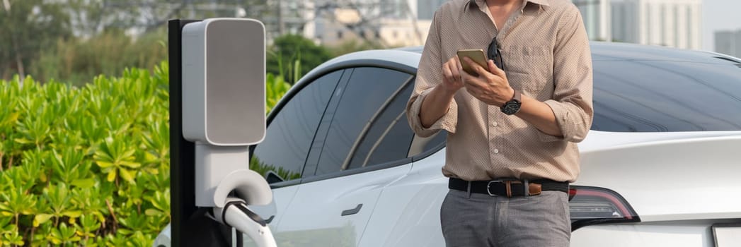 Young man recharge electric car's battery from charging station in outdoor green city park. Rechargeable EV car for sustainable environmental friendly urban travel. Panorama Expedient