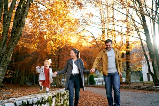 Smiling mom and dad walking with little girl holding hands on the road in the park. High quality photo