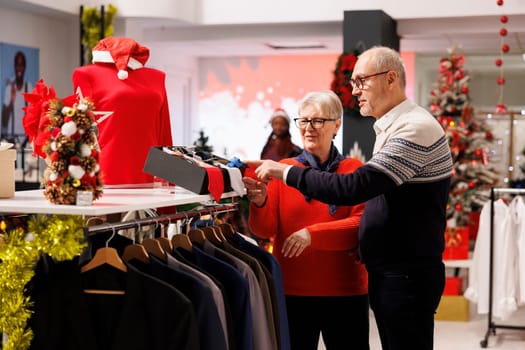 Old customers checking belts and ties in clothing store, looking at accessories to match formal attire for christmas eve dinner outfit. Senior couple searching for clothes during winter sales.