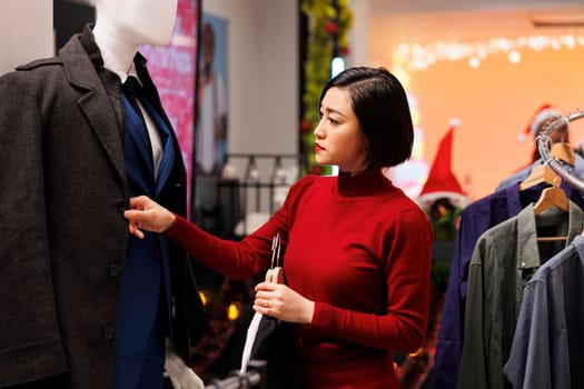 Asian customer browses for clothes on sale, looking for new fashion items in clothing store during christmas seasonal promotions. Woman checking fabric of garments on hangers, buying presents.