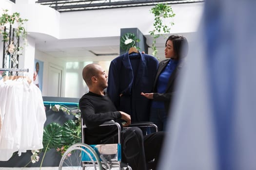 Arab man buyer with disability discussing suit with asian woman seller in department center fashion showroom. Customer in wheelchair examining formal outfit before making purchase