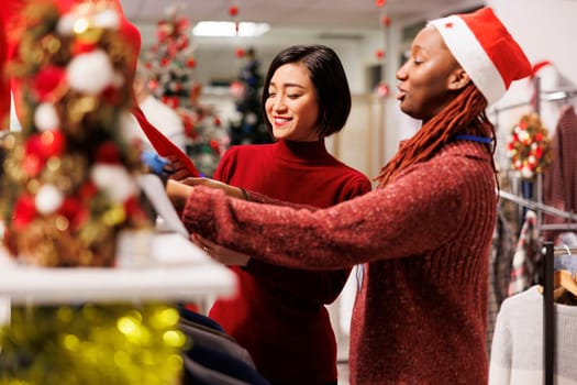 Store clerk helping asian client to find right measurement, recommending clothes and accessories for christmas dinner preparations at mall. Diverse women discussing about fabrics quality.