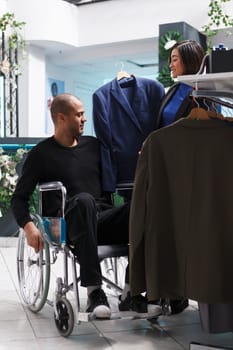 Arab man customer in wheelchair engaging with seller, discussing jackets available in clothing store. Client with disability examining apparel style and size while shopping for formal outfit