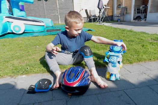 Blonde Caucasian child puts on blue roller skates, sits on the green lawn in the courtyard of the house, early independence, sports life since childhood, growing up as a champion, High quality photo