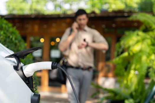 Focused EV electric car recharging at outdoor coffee cafe in springtime garden with blur background of eco friendly man, green city sustainability and environmental friendly EV vehicle. Expedient