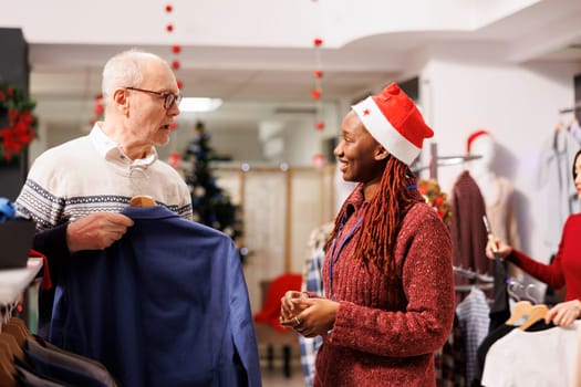 Mall worker showing clothes to client, helping person to choose perfect jacket for festive christmas dinner. African american woman employee recommending clothing for senior customer.