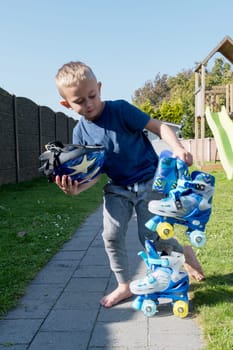 Blonde Caucasian child puts on blue roller skates, sits on the green lawn in the courtyard of the house, early independence, sports life since childhood, growing up as a champion, High quality photo