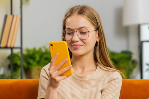 Portrait of woman sitting on sofa uses mobile phone smiling at home living room apartment. Young girl texting share messages content on smartphone social media applications online watching relax movie