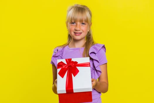 Lovely smiling young preteen child girl kid presenting birthday gift box stretches out hands, offer wrapped present career bonus, celebrating party. Little cute children on studio yellow background