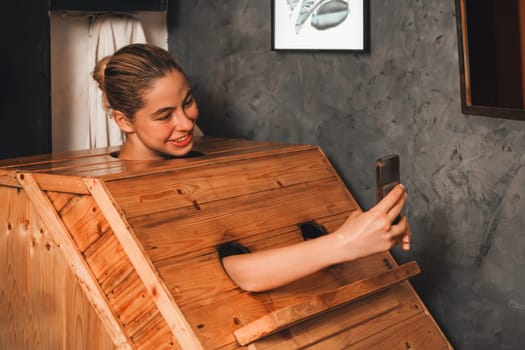 A portrait of gorgeous caucasian woman playing her mobile phone while using wooden sauna cabinet in warm tone. Attractive female with beautiful skin taking a photo. Gray background. Tranquility