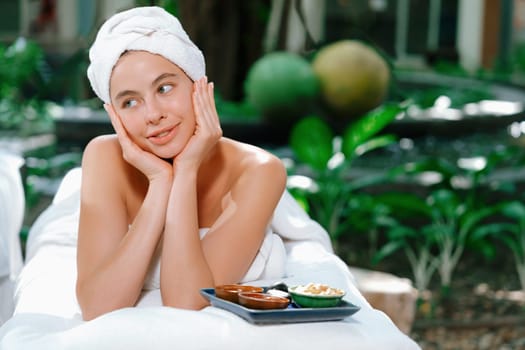 Beautiful caucasian women in white towel smells herbal homemade facial mask while lie on spa bed surrounded by outdoor natural environment. Relaxing and chilling concept. Tranquility.