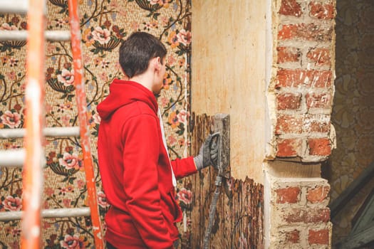 A young caucasian guy in a red body shirt removes retro wallpaper using a steamer machine in an old abandoned house with a broken doorway in the wall, close-up side view.Concept home renovation,interior