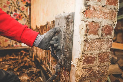 The hands of a young Caucasian man in a red shirt remove retro wallpaper on a steamboat in an old abandoned house with a broken brick doorway, close-up side view. The concept of home renovation, construction work, home.