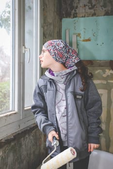 A caucasian teenage girl in a bandana with a paint roller in her hands stands in the bathroom opposite a plastic window and looks with surprise with her mouth open in an old abandoned house, close-up side view.Concept of home renovation,interior of an abandoned house.