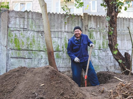 One elderly Caucasian man in a blue uniform with a cap on his head with a happy smile on his face is manually using a shovel to dig the root of an old tree in a deep hole in the backyard of his house on an autumn day, close-up side view. Concept for home renovation, construction work, home.