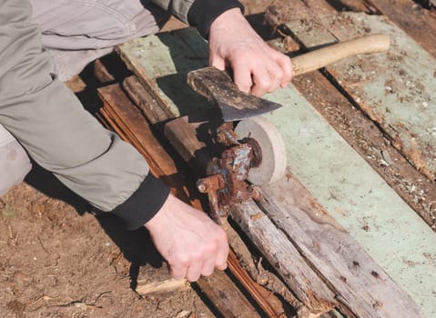 One young unrecognizable Caucasian guy, squatting, manually sharpens an ax with a retro sharpener on a spring day in the backyard of his house on an autumn day, close-up view from above. Concept for home renovation, construction work, home.