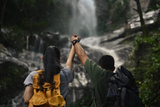 Excited young couple tourist with backpack raising arms enjoying tropical waterfall view.