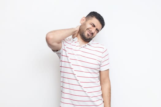Portrait of handsome sick tired bearded man wearing striped t-shirt standing massaging his painful neck, frowning face from terrible pain. Indoor studio shot isolated on gray background.