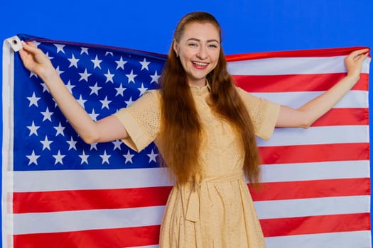 Young adult woman waving and wrapping in American USA flag, celebrating, human rights and freedoms. Independence day. Pretty attractive caucasian girl isolated on blue studio background indoors