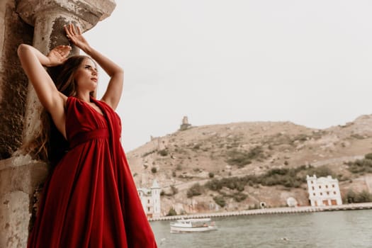 Side view a Young beautiful sensual woman in a red long dress posing on a volcanic rock high above the sea during sunset. Girl on the nature on overcast sky background. Fashion photo