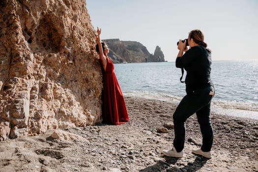 Side view a Young beautiful sensual woman in a red long dress posing on a rock high above the sea during sunrise. Girl on the nature on blue sky background. Fashion photo.