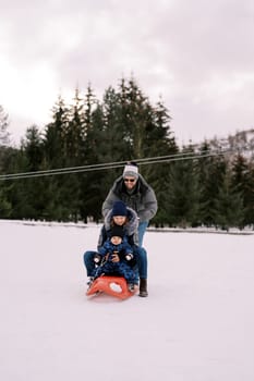 Smiling dad pushes mom with a small child downhill on a sled. High quality photo