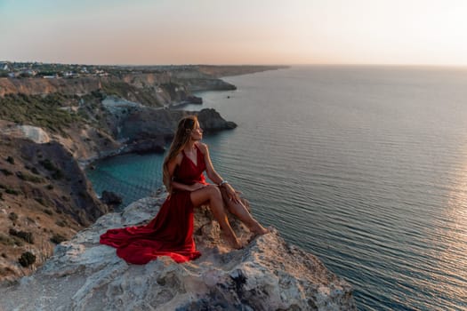 Woman sunset sea red dress, side view a happy beautiful sensual woman in a red long dress posing on a rock high above the sea on sunset