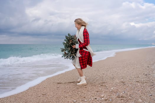 Blond woman holding Christmas tree by the sea. Christmas portrait of a happy woman walking along the beach and holding a Christmas tree in her hands. Dressed in a red coat, white dress