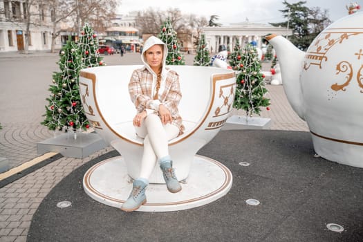 Woman Christmas Square. She sits in a large white cup, dressed in a light suit. With trees decorated with Christmas tinsel in the background.