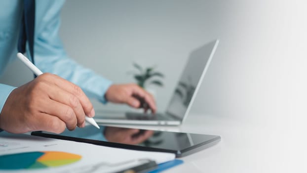 Businessman holding pen pointing at tablet and using computer to work in modern office with copy space.