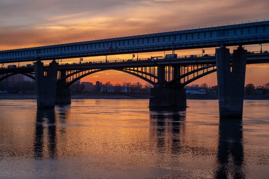 Beautiful view of the bridge over which cars drive at sunset. A river flows under the bridge, reflecting the sunset rays. Bridge in the city of Novosibirsk, Russia