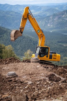 A yellow excavator works in the mountains. Construction of roads and houses in beautiful mountains.